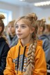 Young blonde girl with long braid wearing hoodie with x-ray of human chest and ribs print sits in audience hall with other pupils.
