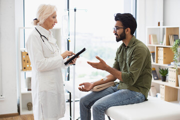 Wall Mural - Medical specialist adjusts composition of drugs looking at electronic history of patient in exam room. Bearded man receiving chemotherapy looking at nurse with tablet examining condition of patient.