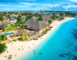 Aerial view of sandy beach, blue sea, bungalows, green palms, umbrellas, swimming people at sunset. Summer travel in Nungwi, Zanzibar island. Tropical landscape. Top drone view of luxury resort. Ocean