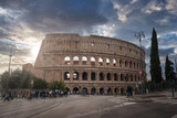 Fototapeta  - Iconic ancient amphitheater in Rome, Italy shows Colosseum's grandeur with multiple arches and historical architecture. Overcast sky, modern elements, tourists, pedestrians nearby.