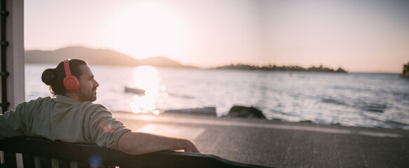 Portrait of a young man in bright large headphones listening to music, audiobook by the sea at sunset.