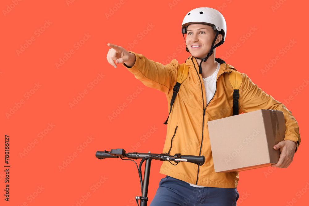 Male courier with cardboard box and kick scooter pointing at something on orange background