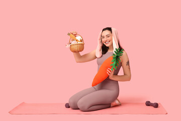 Poster - Happy female fitness coach in bunny ears with Easter basket, toy carrot and dumbbells on pink background