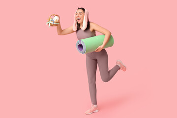 Poster - Happy female fitness coach in bunny ears with Easter basket and yoga mat on pink background