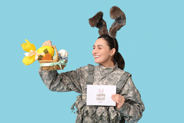 Canvas Print - Happy female soldier in bunny ears with greeting card, Easter basket and chocolate egg on blue background