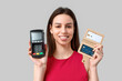 Young woman with credit card holder and payment terminal on grey background
