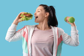 Poster - Sporty young woman with sandwich and dumbbell on blue background. Overeating concept