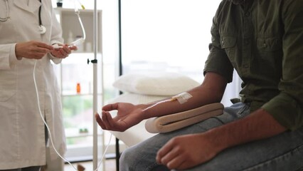 Wall Mural - Young bearded man in casual wear taking dropper while mature female making system review. Efficient general practitioner checking solution for chemotherapy of male sitting on exam couch.