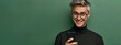 Photo of An elegant man in his 20s with short hair, smiling while using an smartphone mobile phone on a green background with copy space for text or product. Web banner with copyspace
