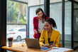 © apichat - A businesswoman in a yellow blazer engaging in a productive discussion with a colleague in a red jacket over work documents in an office.
