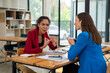 © apichat - Two professional women in a lively discussion at an office desk, with one in a yellow blazer gesturing and the other showing surprise.