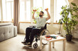 Young man sitting in wheelchair in headphones, training, doing exercises with dumbbells at home. Staying active at home. Concept of healthcare, lifestyle, wellness, comfort, empowerment