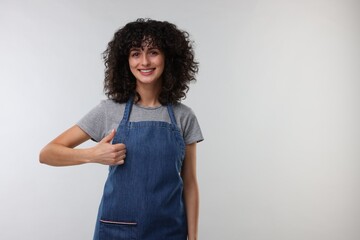 Wall Mural - Happy woman wearing kitchen apron and showing thumbs up on light grey background, space for text. Mockup for design