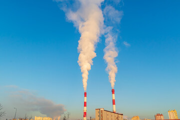 Two tall red and white smoke stacks are spewing smoke into the sky. The sky is clear and blue, with no clouds in sight. The smoke is billowing high into the air, creating a dramatic and ominous scene