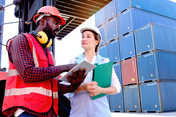 Two workers with safety helmet at logistic shipping cargo containers yard. African engineer man using digital tablet to report results to beautiful woman boss who holds document folder at workplace.