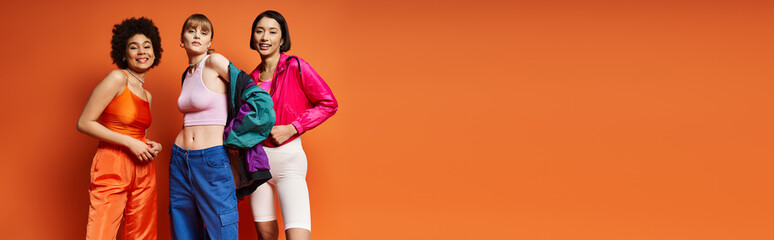 Three women of different ethnic backgrounds standing together in front of an orange studio background.