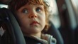 A young child with curly hair wearing a car seat looking out of a car window with a curious expression.