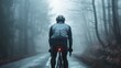 A lone cyclist in a gray jacket and black helmet riding a red-lit bicycle pedaling down a foggy tree-lined road.