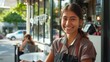 Smiling young woman in apron standing outside a coffee shop with a warm and inviting atmosphere.