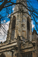 Gothic church tower with intricate stone details, framed by bare tree branches against a blue sky with fluffy clouds in York, North Yorkshire, England.