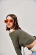 A stylish brunette woman standing confidently in a green top and trendy orange sunglasses.