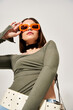 A young woman with brunette hair posing in a studio setting wearing a vibrant green top and trendy orange sunglasses.