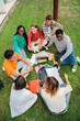 © Jose Calsina - Vertical. Multiracial students using a laptop computert sitting in a circle on the grass, doing homework outside at university campus. Young high school people working on a academy task or schoolwork