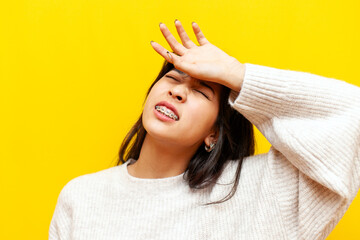 young tired asian woman with braces suffers and is sad on a yellow isolated background, korean unhappy girl wipes her forehead with her hand and shows dissatisfaction