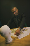 Fototapeta Tęcza - An African-American man in a shirt and tie is strangling the head of a mannequin. There is money, papers, documents on the table. Concept business, competition, stress