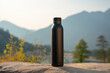 Black water bottle mockup placed on rock with mountains in background. Mock up of blank reusable set against the tranquil beauty of lake