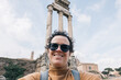 A  woman with sunglasses and backpack is taking a selfie in the Roman forum, she is smiling and look at camera