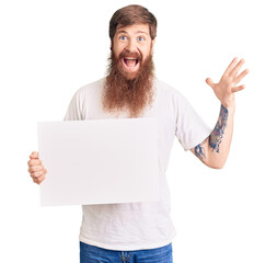 Canvas Print - Handsome young red head man with long beard holding blank empty banner celebrating victory with happy smile and winner expression with raised hands