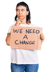 Sticker - Young woman holding we need a change banner serious face thinking about question with hand on chin, thoughtful about confusing idea