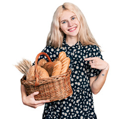 Canvas Print - Young caucasian woman holding wicker basket with bread smiling happy pointing with hand and finger