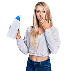 Canvas Print - Beautiful young blonde woman holding detergent bottle covering mouth with hand, shocked and afraid for mistake. surprised expression