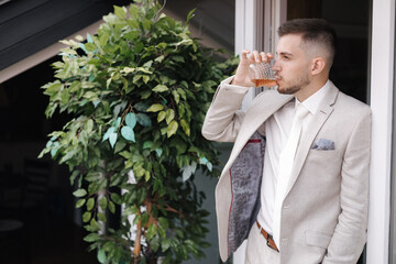 Canvas Print - Portrait of young and handsome groom in light colour suit hold glass of whiskey and drink. Side view. Place for text