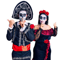 Wall Mural - Young couple wearing mexican day of the dead costume over background asking to be quiet with finger on lips pointing with hand to the side. silence and secret concept.