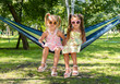 Two adorable five-year-old sisters are laughing and enjoying a peaceful moment as sitting together in a hammock, surrounded by lush greenery in a sunny summer park.