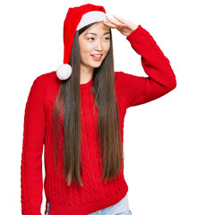 Canvas Print - Young chinese woman wearing christmas hat very happy and smiling looking far away with hand over head. searching concept.