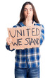 Young adult man with long hair holding united we stand banner covering mouth with hand, shocked and afraid for mistake. surprised expression
