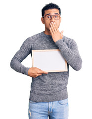 Sticker - Hispanic handsome young man holding blank empty banner covering mouth with hand, shocked and afraid for mistake. surprised expression