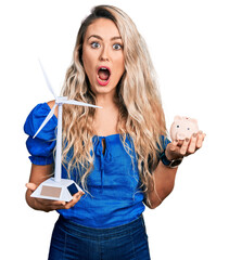 Sticker - Young blonde woman holding windmill and piggy bank afraid and shocked with surprise and amazed expression, fear and excited face.