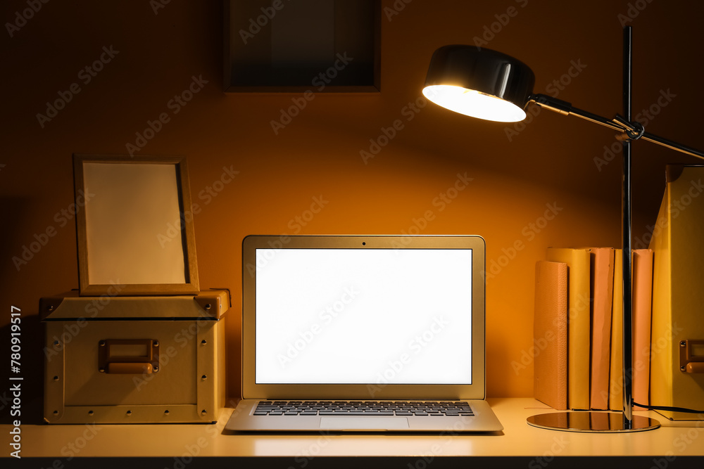 Comfortable workplace with glowing lamp and modern laptop near orange wall