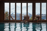 Fototapeta  - Indoor swimming pool with wooden interior and glass doors showing snow covered mountains. Relaxing setting, potted plants, balcony with outdoor furniture. Serene ambiance. Ideal for relaxation.