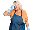 Middle age blonde woman wearing cleaner apron and gloves tired rubbing nose and eyes feeling fatigue and headache. stress and frustration concept.