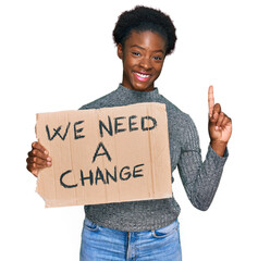 Wall Mural - Young african american girl holding we need a change banner surprised with an idea or question pointing finger with happy face, number one