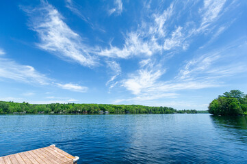 Wall Mural - A tranquil lakeside scene unfolds, with quaint cottages nestled amidst lush greenery on the opposite shore. A wooden dock peeks into view from the bottom left corner.