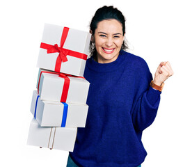 Young hispanic woman holding gifts screaming proud, celebrating victory and success very excited with raised arm