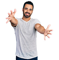 Sticker - Young hispanic man with beard wearing casual grey t shirt looking at the camera smiling with open arms for hug. cheerful expression embracing happiness.