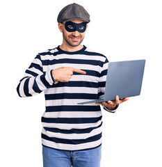 Canvas Print - Young handsome man wearing burglar mask using laptop smiling happy pointing with hand and finger